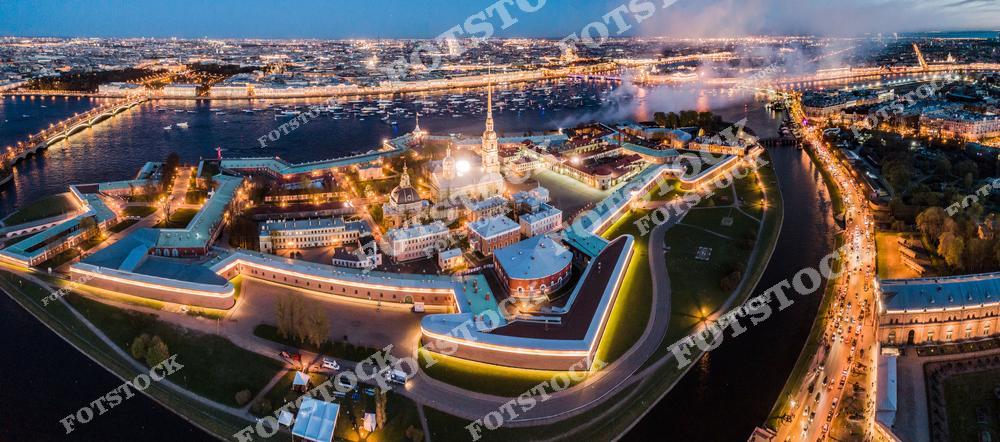 Панорамная фотография Петропавловской крепости и центра Санкт-Петербурга с высоты птичьего полета вечером. На снимке видны крепостные стены, собор Петра и Павла, река Нева, разводные мосты и огни города. Съемка с дрона позволяет оценить масштабы архитектурного ансамбля, панораму набережных и величие Северной столицы России. Фотография передает атмосферу вечернего Петербурга, торжественность исторического центра и красоту городских огней. Снимок идеально подходит для туристических публикаций, фотостоков, рекламы, оформления интерьеров и материалов о достопримечательностях города.